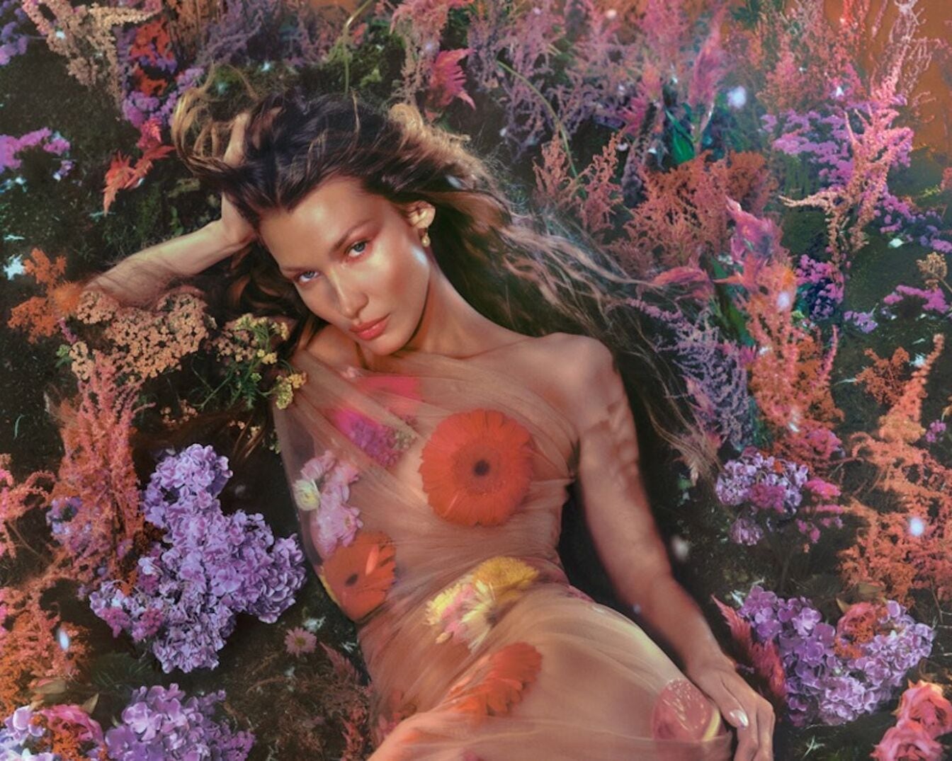 Une femme aux longs cheveux ondulés est allongée parmi des fleurs violettes, orange et roses, portant une robe transparente ornée de fleurs.