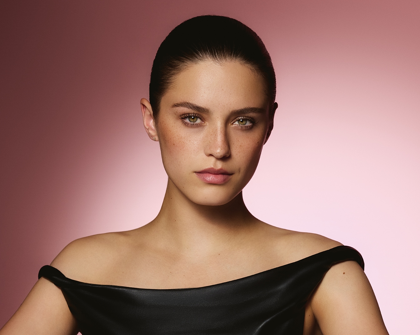 Portrait d'une jeune femme aux cheveux bruns tirés en arrière, portant une robe noire à épaules dénudées, avec un fond rose dégradé.