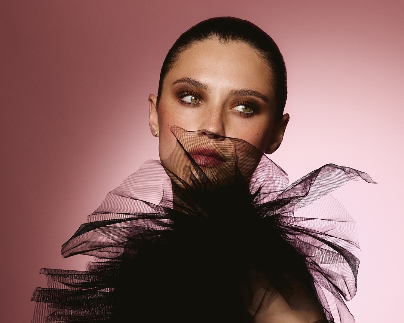 Portrait d'une femme avec un voile noir en tulle, regardant vers la droite, sur un fond rose uni, capturant une esthétique à la fois mystérieuse et élégante.