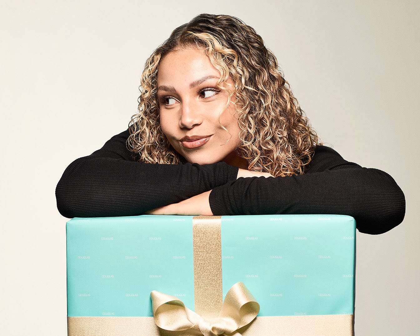 Una donna appoggiata a un regalo avvolto in carta azzurra con nastro dorato, che mostra un'espressione pensierosa.