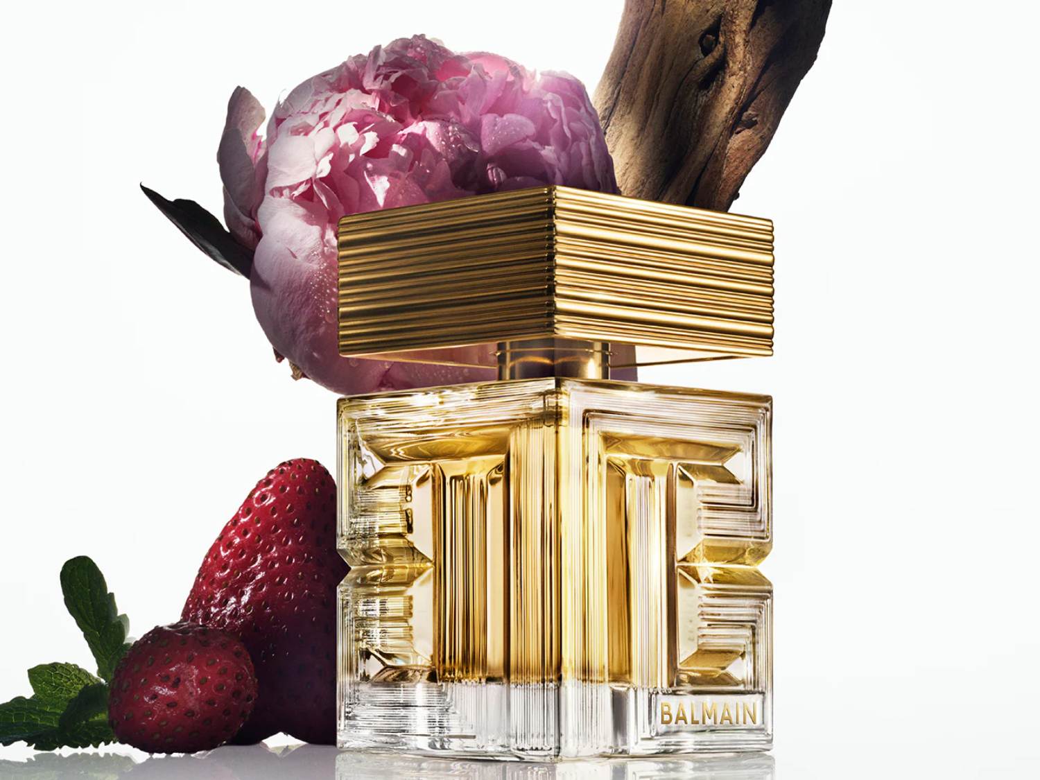 Elegancki flakon perfum Balmain w złotym odcieniu, ozdobiony kwiatem piwonii, truskawkami i gałązką, tworząc luksusową kompozycję.