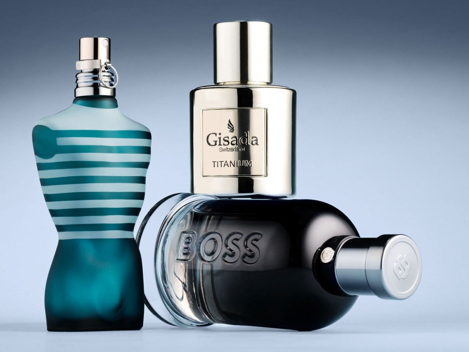 Ecco tre flaconi di profumo: uno a forma di busto maschile a righe bianche e blu, uno dorato di Gisada Switzerland Titanium e uno nero orizzontale di Boss.