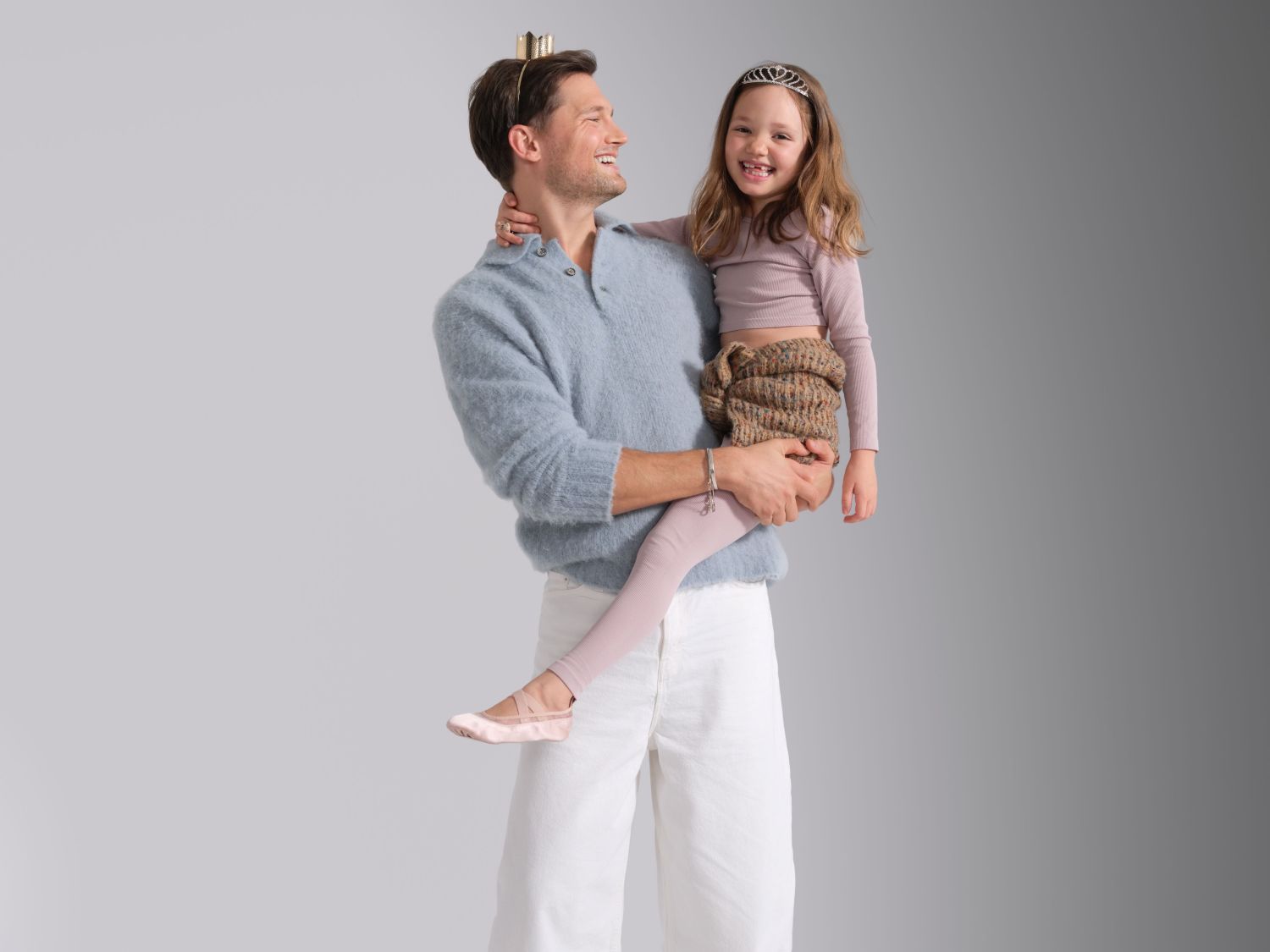 Un uomo sorridente con una corona tiene in braccio una bambina sorridente con un diadema, entrambi vestiti in modo casual ed elegante.
