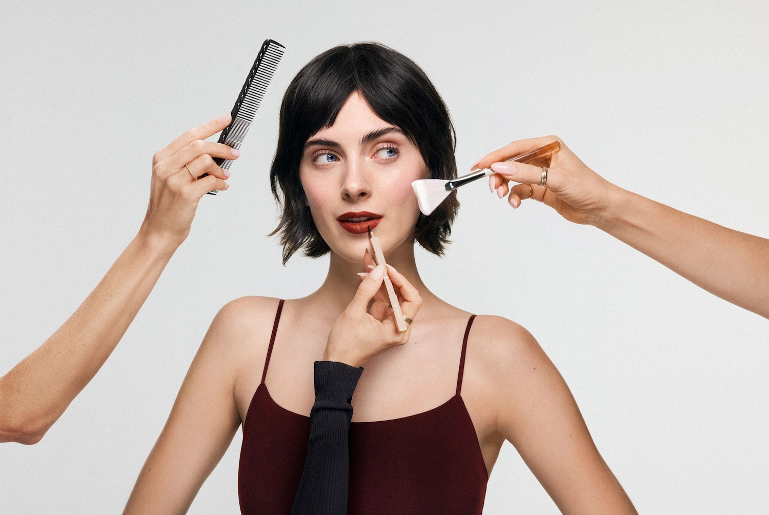 Une femme aux cheveux bruns et aux yeux bleus se fait maquiller les lèvres avec un crayon à lèvres marron, tandis qu'une main tient un peigne noir et une autre applique du blush avec un pinceau.