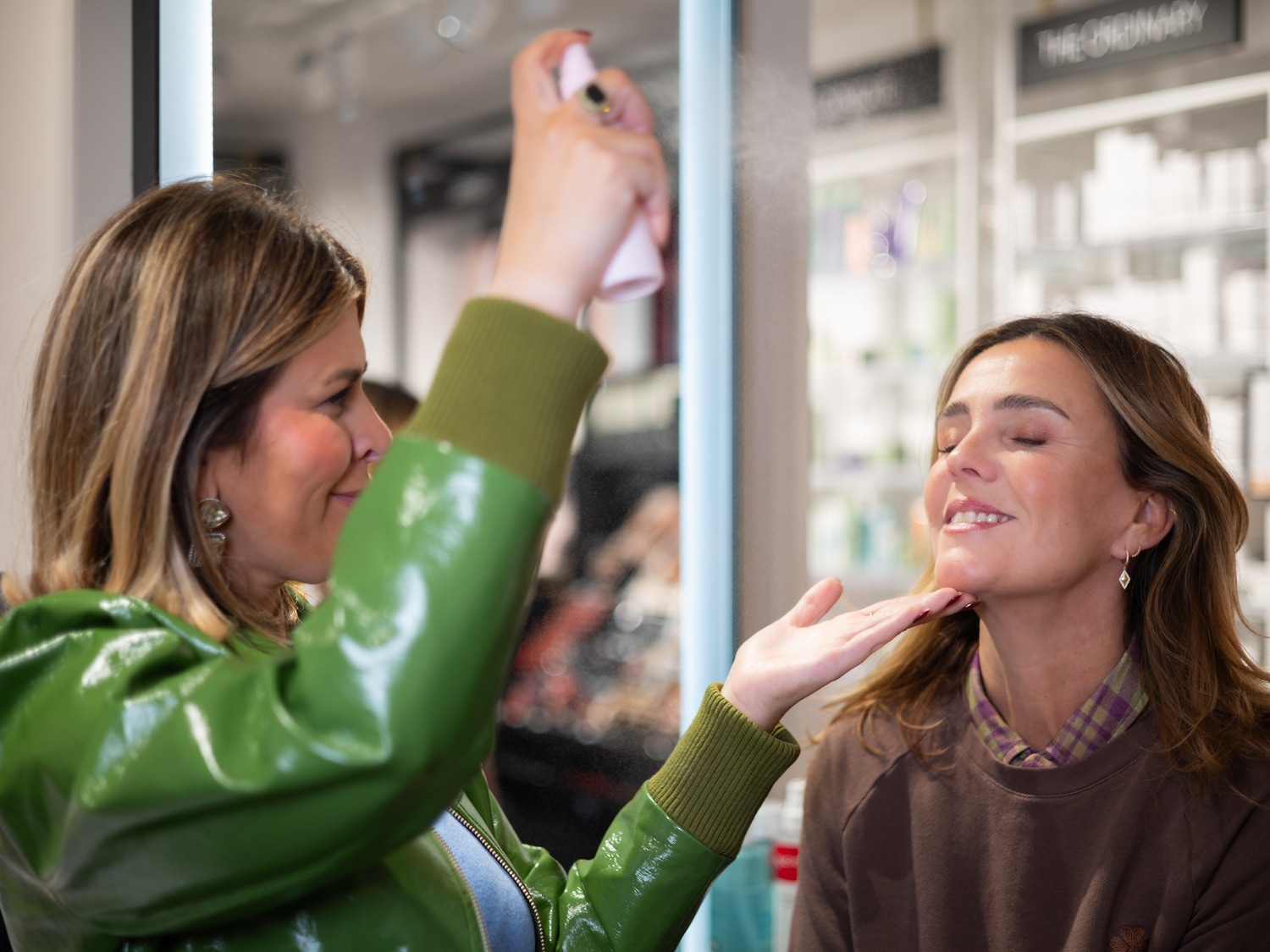 Une femme en veste verte applique un spray sur le visage d'une autre femme aux yeux clos, souriant légèrement.