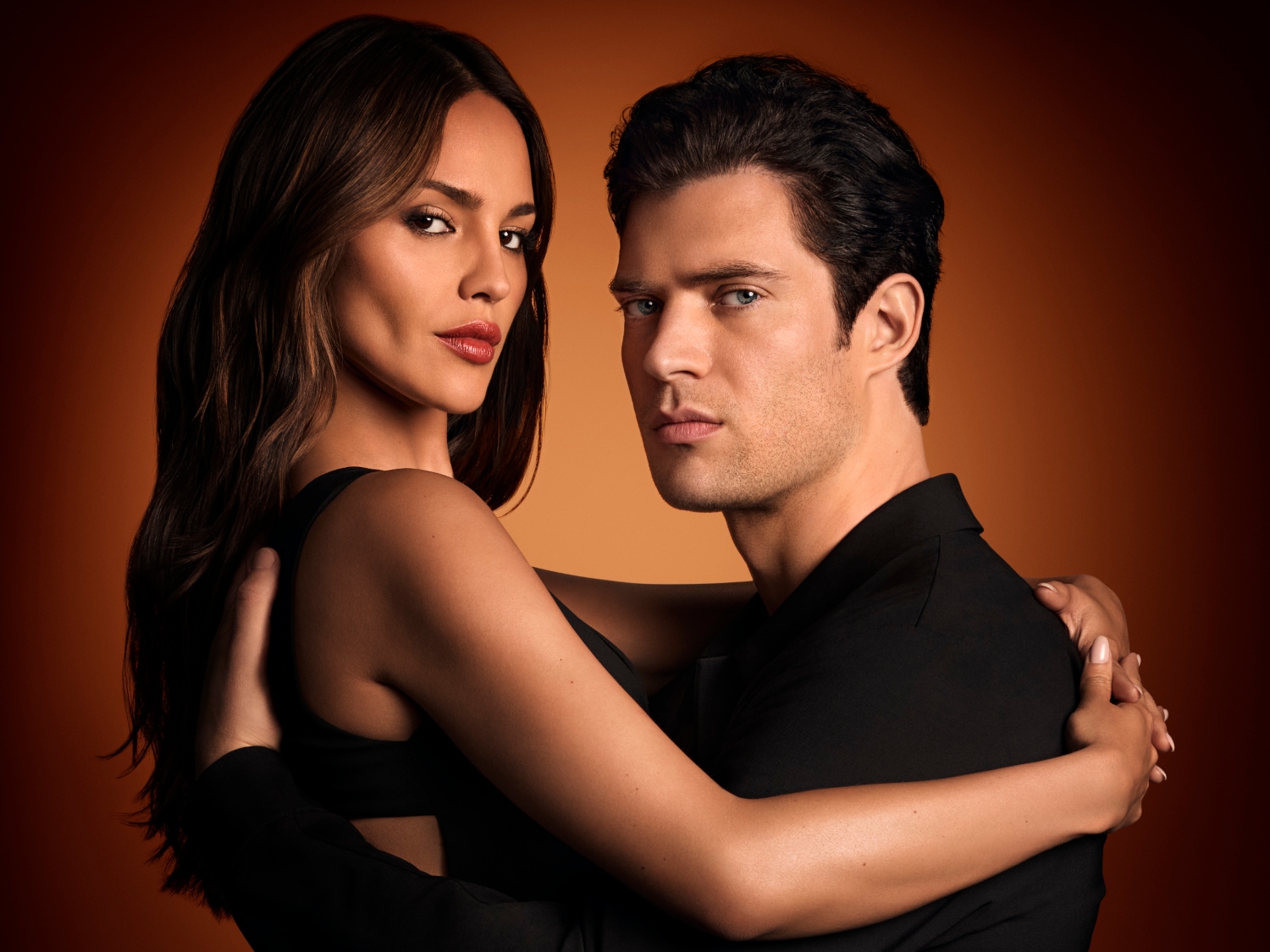 Na tle w odcieniach brązu, kobieta z długimi brązowymi włosami i mężczyzna w czarnej koszuli obejmują się, patrząc w obiektyw.