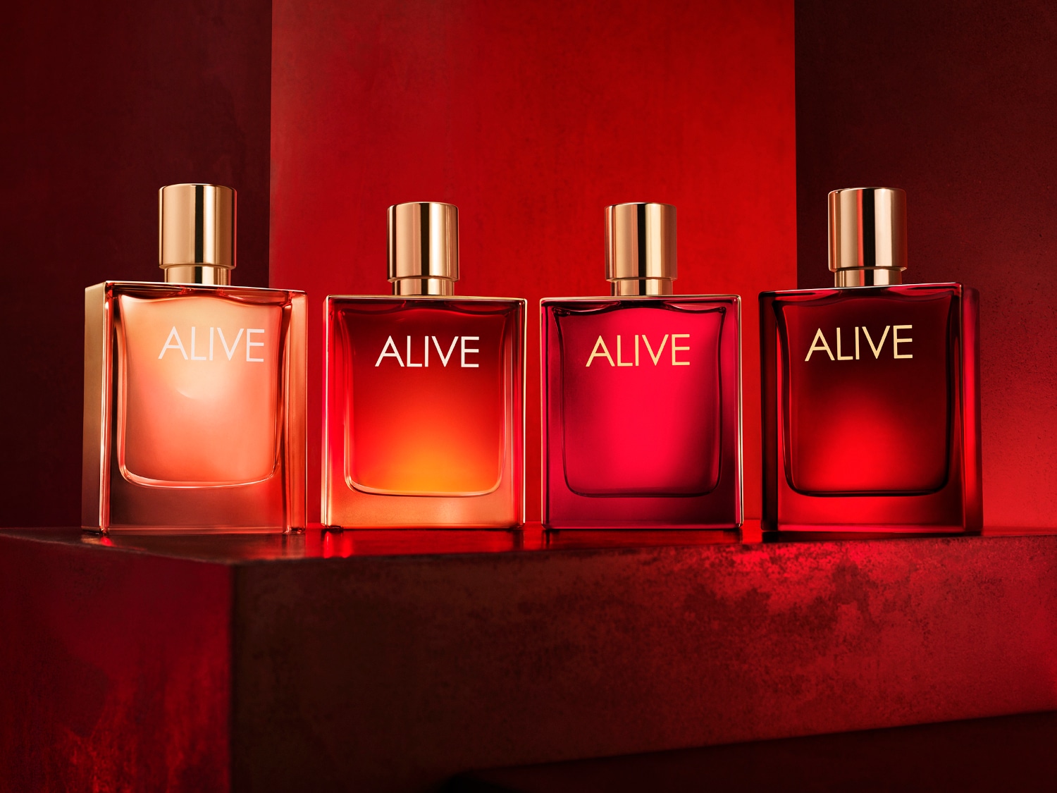 Cztery flakony perfum Alive w odcieniach czerwieni, od jasnego do ciemnego, z złotymi korkami, ustawione na czerwonym tle, emanujące ciepłem i energią.