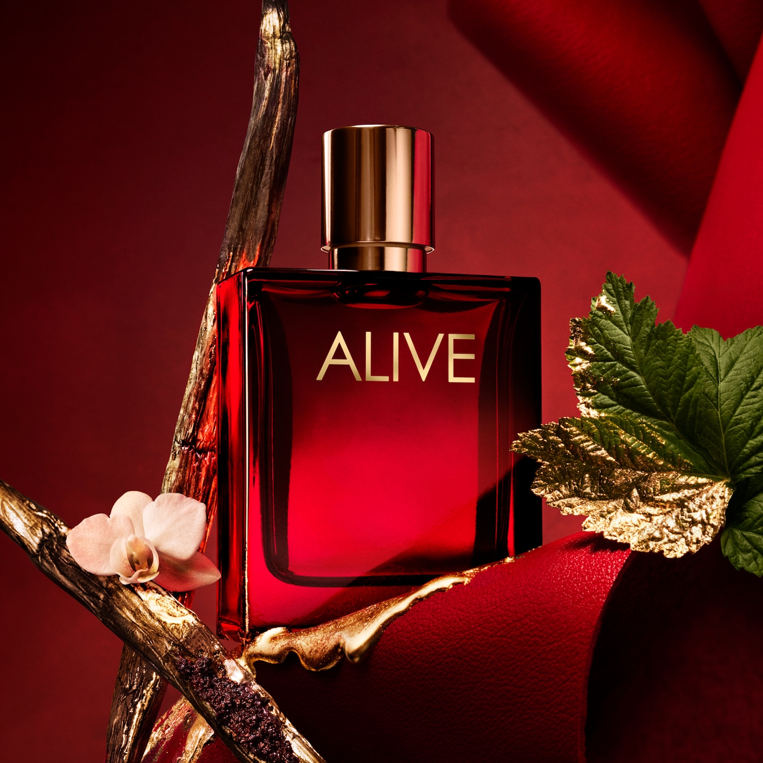 Czerwony flakon perfum ALIVE z złotymi akcentami, otoczony dekoracyjnymi elementami, takimi jak złota gałązka, liść i delikatny kwiat.