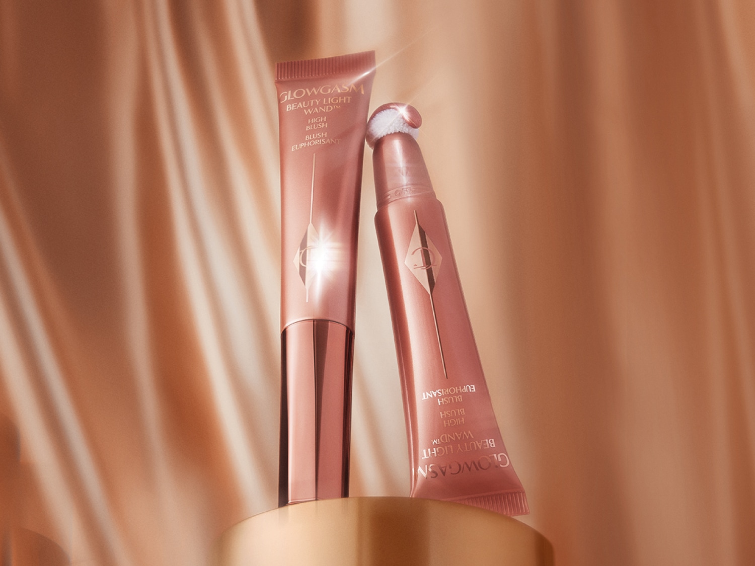 Auf einem goldenen Sockel stehen zwei Glowgasm Beauty Light Wand High Blush Tuben in Roségold, die einen strahlenden Teint versprechen.