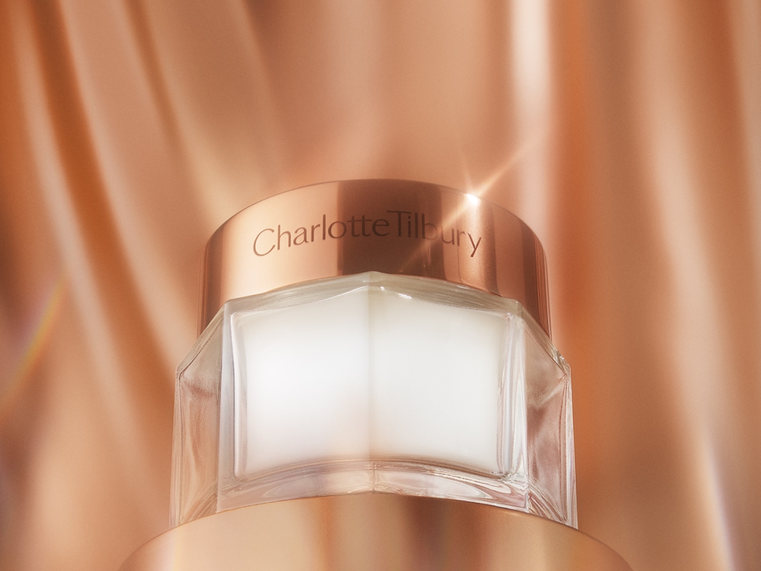 Nahaufnahme eines Charlotte Tilbury Tiegel mit goldfarbenem Deckel und klarer Basis, gefüllt mit einer weißen Creme, vor einem schimmernden, pfirsichfarbenen Hintergrund.
