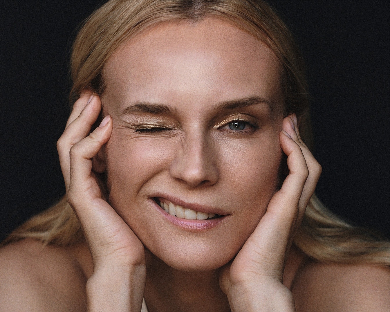 Portrait d'une femme blonde souriante avec un fard à paupières doré scintillant sur une paupière, les mains sur les joues, sur un fond noir uni.