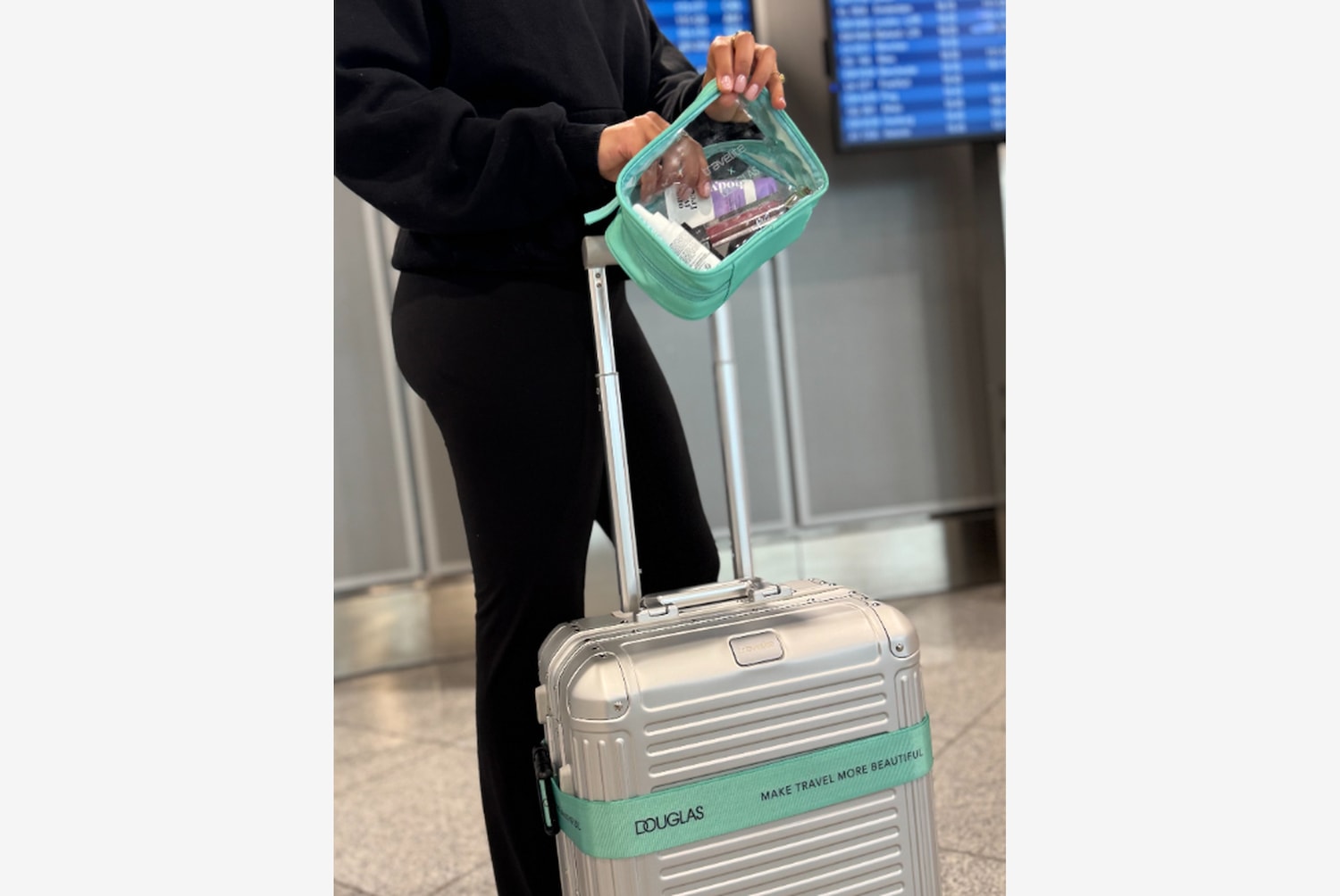 Eine Person mit schwarzer Kleidung hält eine durchsichtige Kosmetiktasche mit mintgrünen Akzenten über einem silbernen Rollkoffer mit einem mintgrünen Band, das mit "Douglas Make travel more beautiful" beschriftet ist.