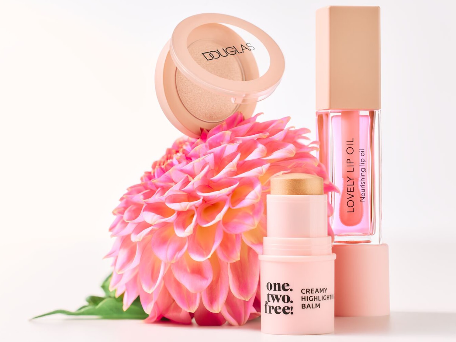 Een close-up van Douglas make-up producten, waaronder een highlighter, lipolie en creamy highlighting balm, geplaatst voor een roze bloem.