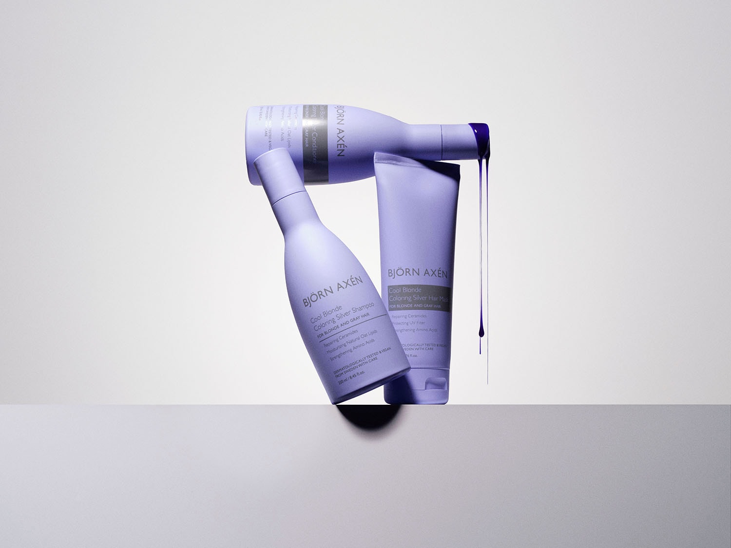 Fioletowe opakowania szamponu, odżywki i maski do włosów Björn Axén Cool Blonde Coloring Silver, przeznaczone do włosów blond i siwych, obok których widać wyciekający fioletowy płyn.