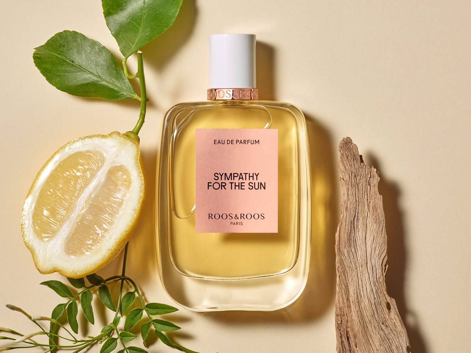 Un flacon de parfum transparent "Sympathy for the Sun" de Roos&Roos Paris, avec une étiquette rose, est entouré d'une demi-citron, d'une branche verte et d'un morceau de bois sur un fond beige.