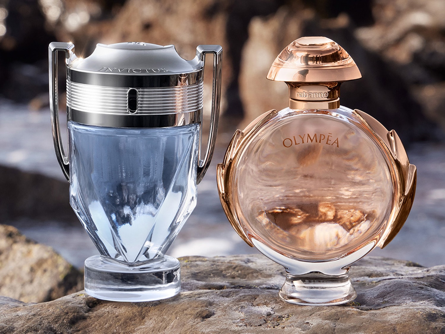 Dva flakony parfémů Paco Rabanne, Invictus s modrou tekutinou ve stříbrné trofeji a Olympéa s růžovou tekutinou v kruhové zlaté nádobě.