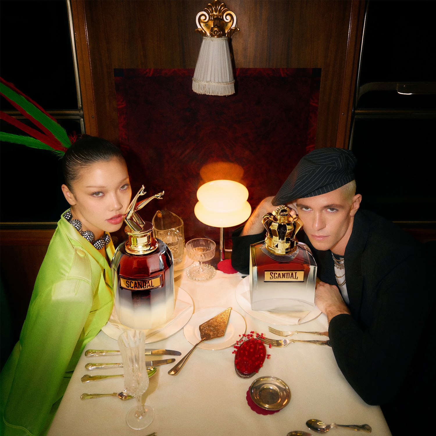 Deux mannequins posent de chaque côté de la table avec les parfums Scandal, l'un avec un flacon rouge couronné d'une figurine dorée, l'autre avec un flacon transparent surmonté d'une couronne dorée.
