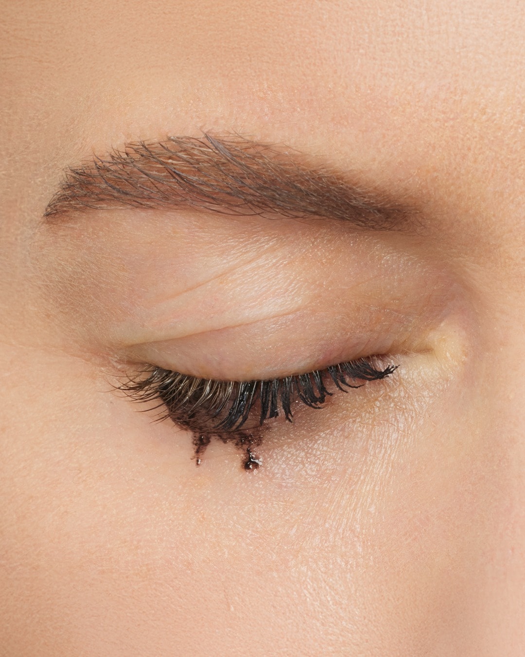 Close-up van een gesloten oog met lange, donkere wimpers waar mascara van afloopt, waardoor er kleine zwarte druppels op de huid eronder ontstaan.