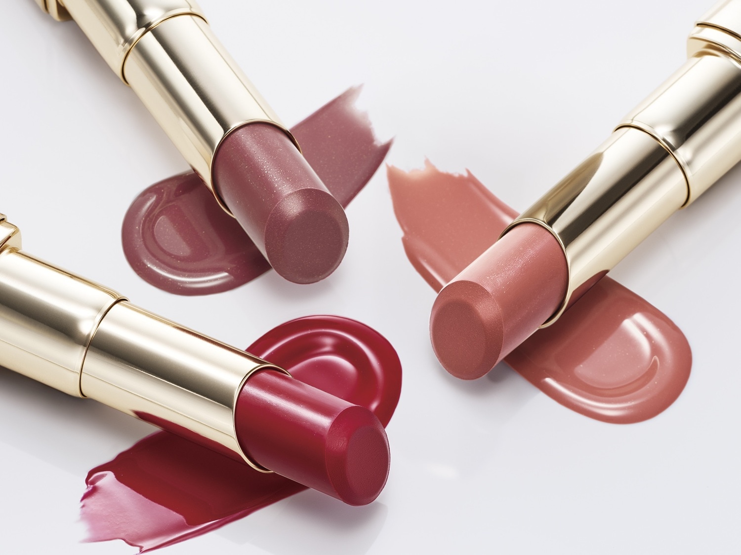 Trzy szminki w złotych opakowaniach leżą obok próbek w kolorach: czerwonym, różowym i fioletowym.
