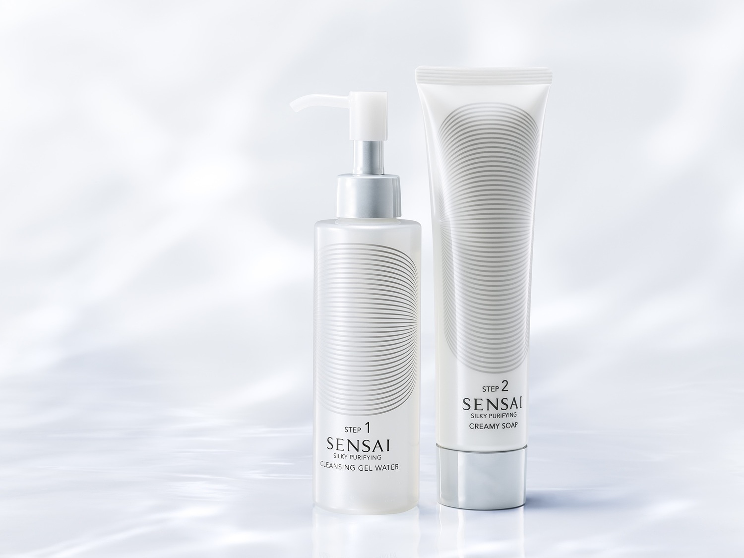 Dwa białe opakowania produktów do oczyszczania twarzy Sensai Silky Purifying: żel do demakijażu w butelce z pompką (krok 1) i kremowe mydło w tubie (krok 2).