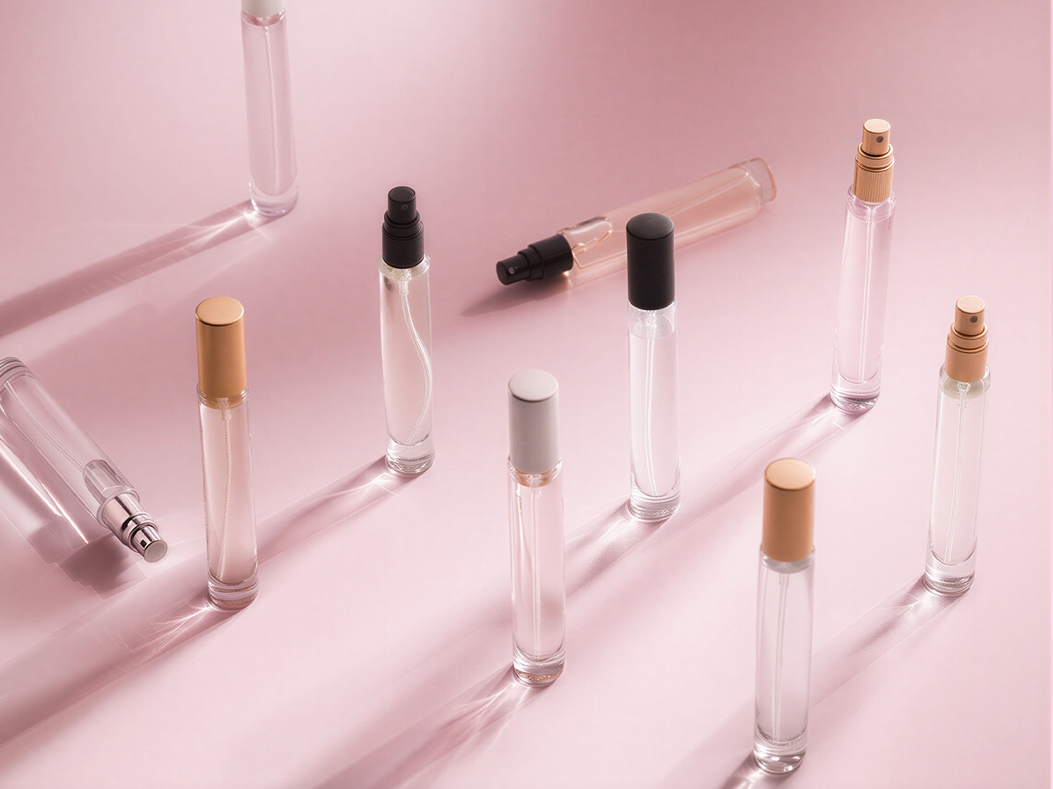Découvrez une collection de vaporisateurs de parfum élégants, présentés dans des flacons transparents aux bouchons dorés, noirs et argentés, sur un fond rose doux.