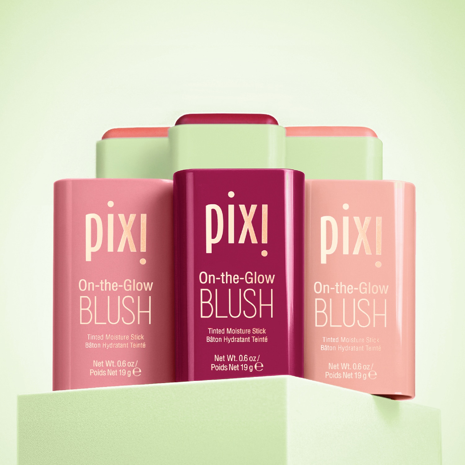Drie Pixi On-the-Glow Blush sticks in roze, framboos en perzik tinten, gestapeld op een lichtgroene achtergrond, voor een getinte, hydraterende kleur.