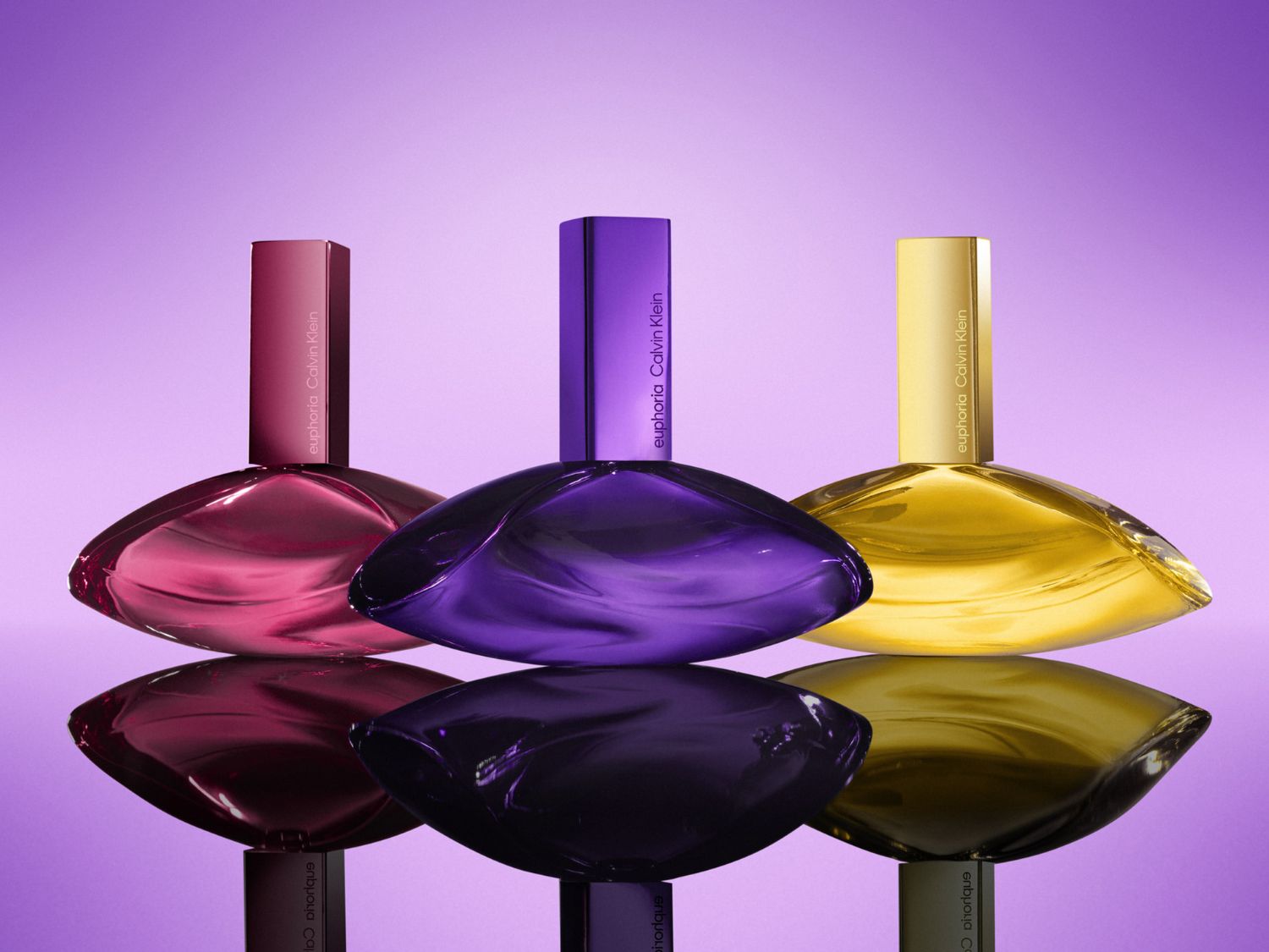 Tri fľaštičky Calvin Klein Euphoria, každá v inej farbe - ružová, fialová a zlatá - s hladkým, lesklým povrchom, ktoré odrážajú svetlo na fialovom pozadí.
