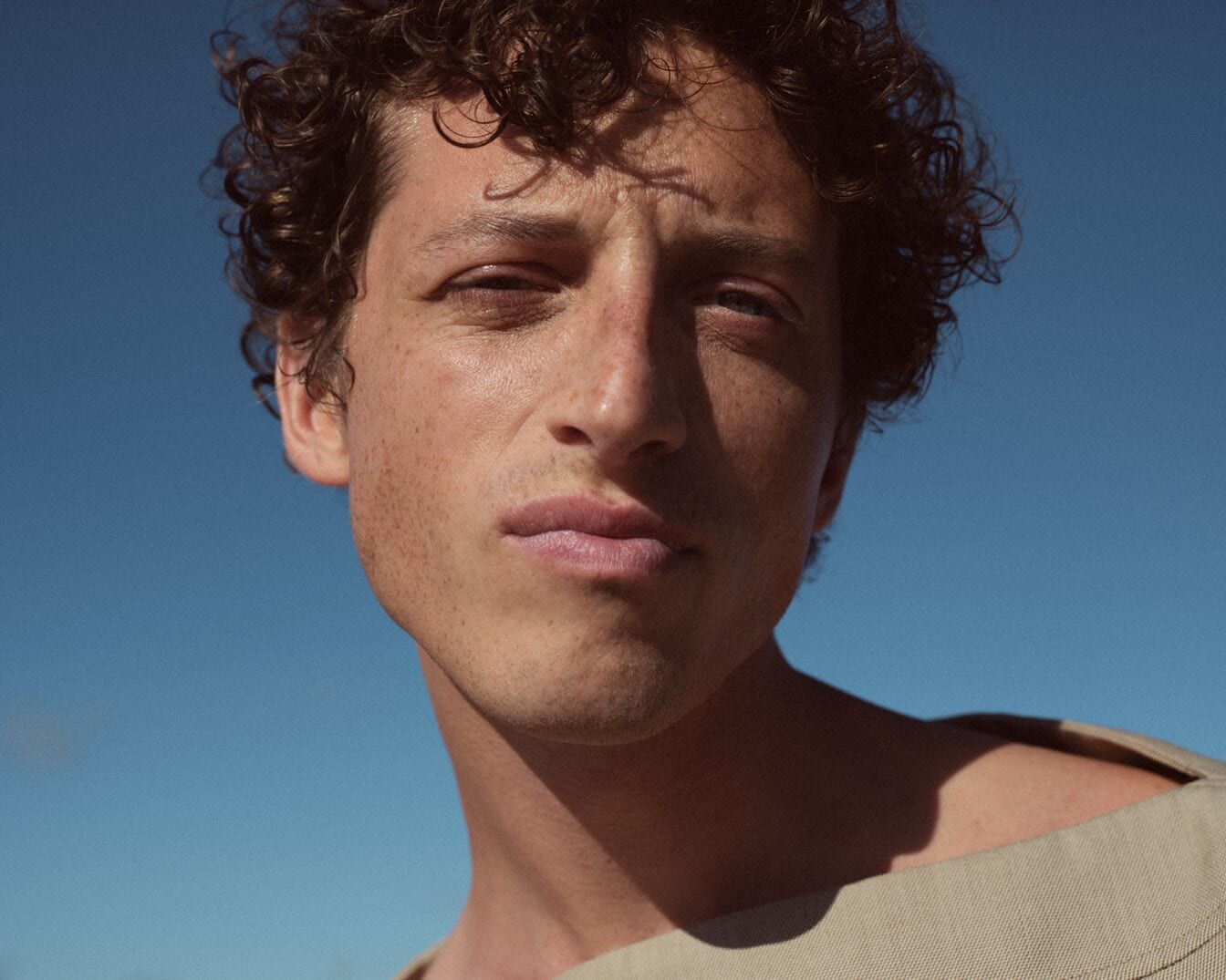 Nahaufnahme eines Mannes mit lockigem, braunem Haar, Sommersprossen und einem beigen Oberteil vor einem strahlend blauen Himmel.