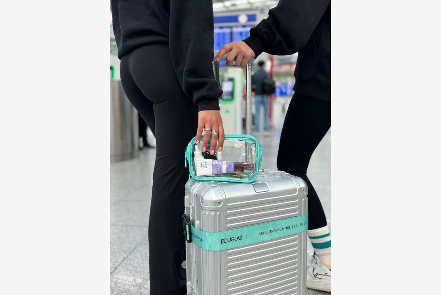 Silberner Rollkoffer mit türkisfarbenem Band mit der Aufschrift "MAKE TRAVEL MORE BEAUTIFUL" und durchsichtiger Kosmetiktasche mit verschiedenen Beauty-Produkten.