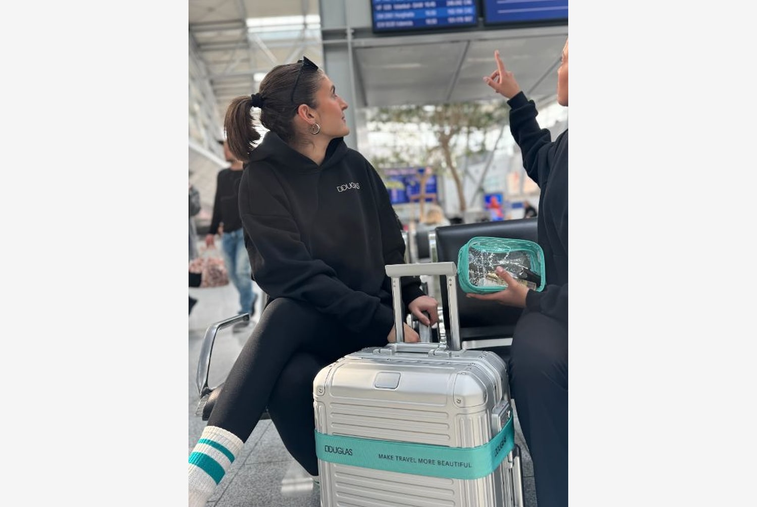 Eine Frau mit einem schwarzen Douglas-Hoodie sitzt mit einem silbernen Koffer mit der Aufschrift "Make travel more beautiful" in einem Wartebereich.