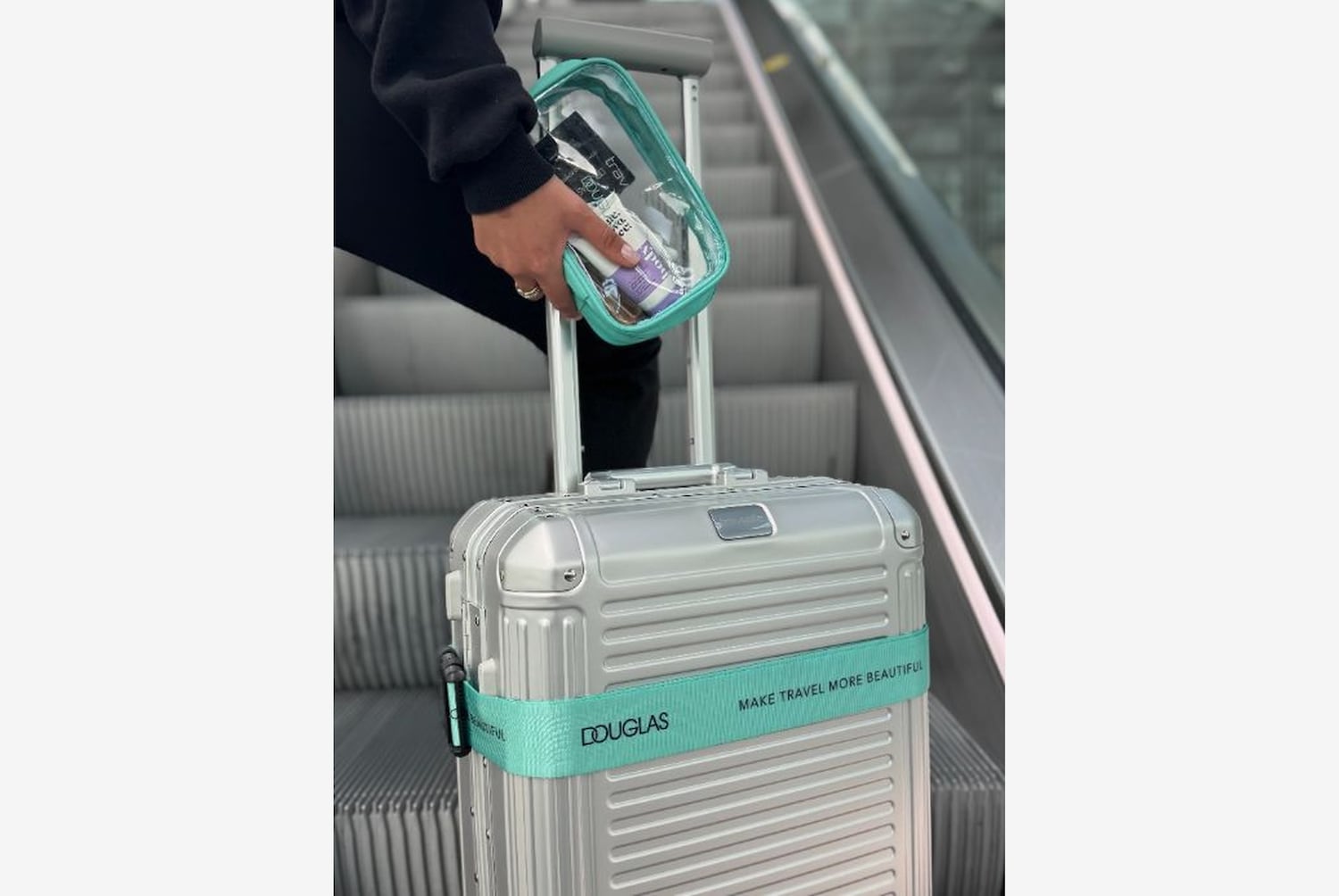 Eine Person hält eine durchsichtige Tasche mit Toilettenartikeln über einem silbernen Rollkoffer mit einem türkisfarbenen Band, auf dem „MAKE TRAVEL MORE BEAUTIFUL“ steht.