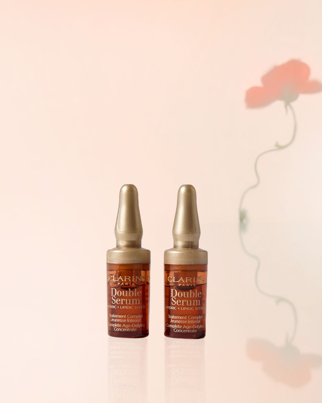 Zwei Flaschen Clarins Double Serum mit goldfarbenen Verschlüssen stehen vor einem zarten Hintergrund mit Blumenschatten, ein komplettes Anti-Aging-Konzentrat für intensive Jugendlichkeit.