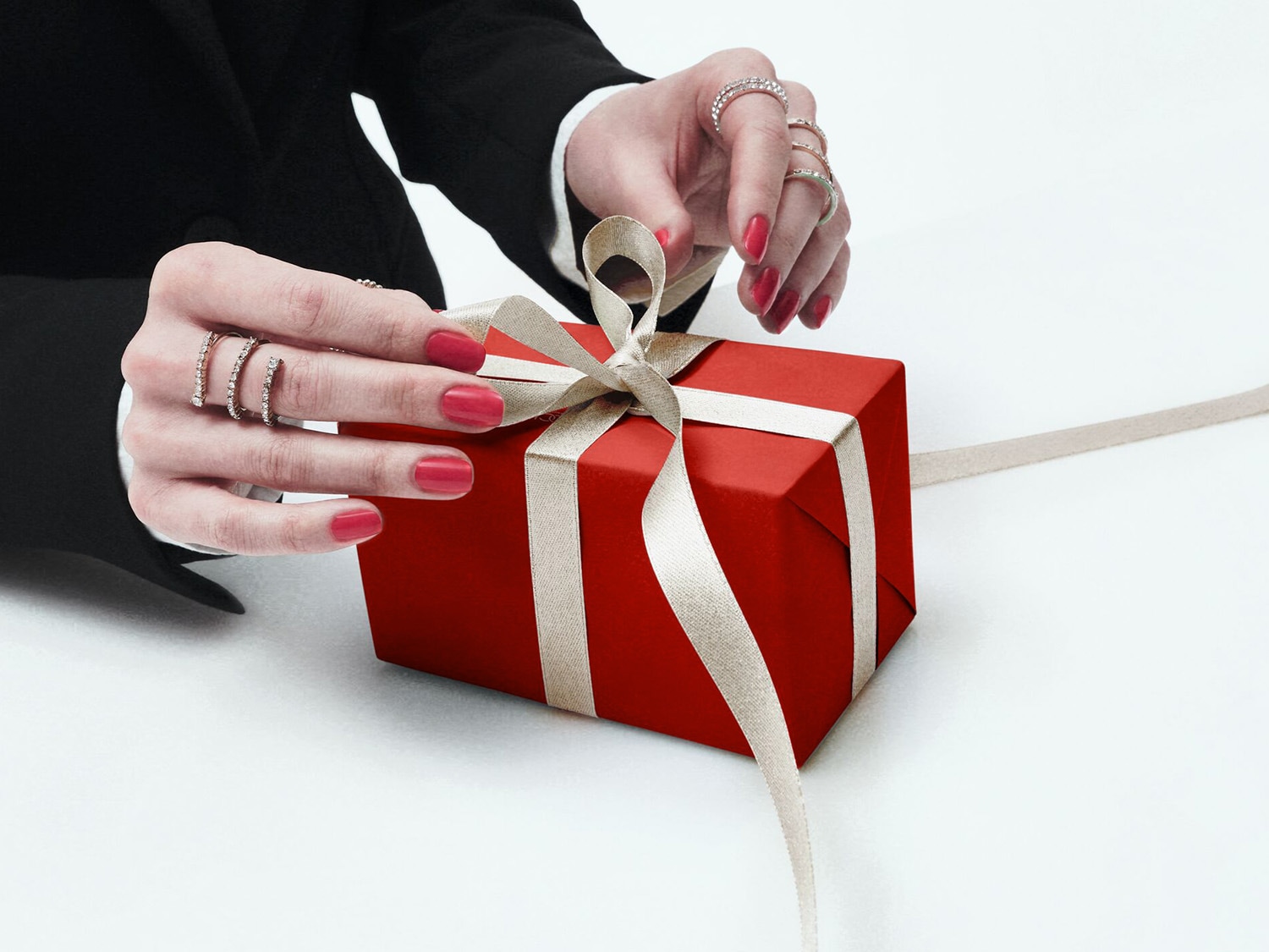 Gros plan sur les mains d'une personne parée de bagues, dénouant délicatement le ruban d'un cadeau emballé dans un papier rouge vif.