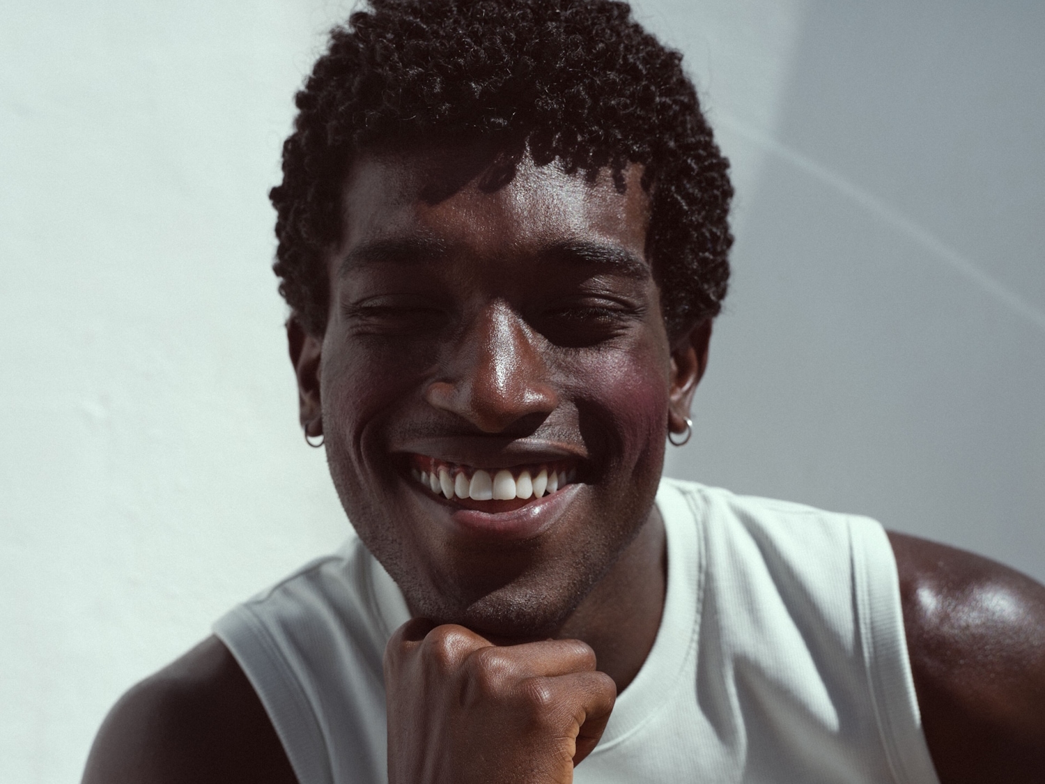 Ein lächelnder, dunkelhäutiger Mann mit krausem Haar, der ein weißes Tanktop trägt und einen silbernen Ohrring trägt, strahlt vor einer hellen Wand.