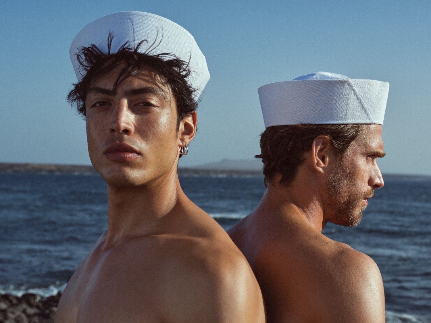 Zwei Männer mit freiem Oberkörper und weißen Matrosenhüten stehen vor dem blauen Meer.