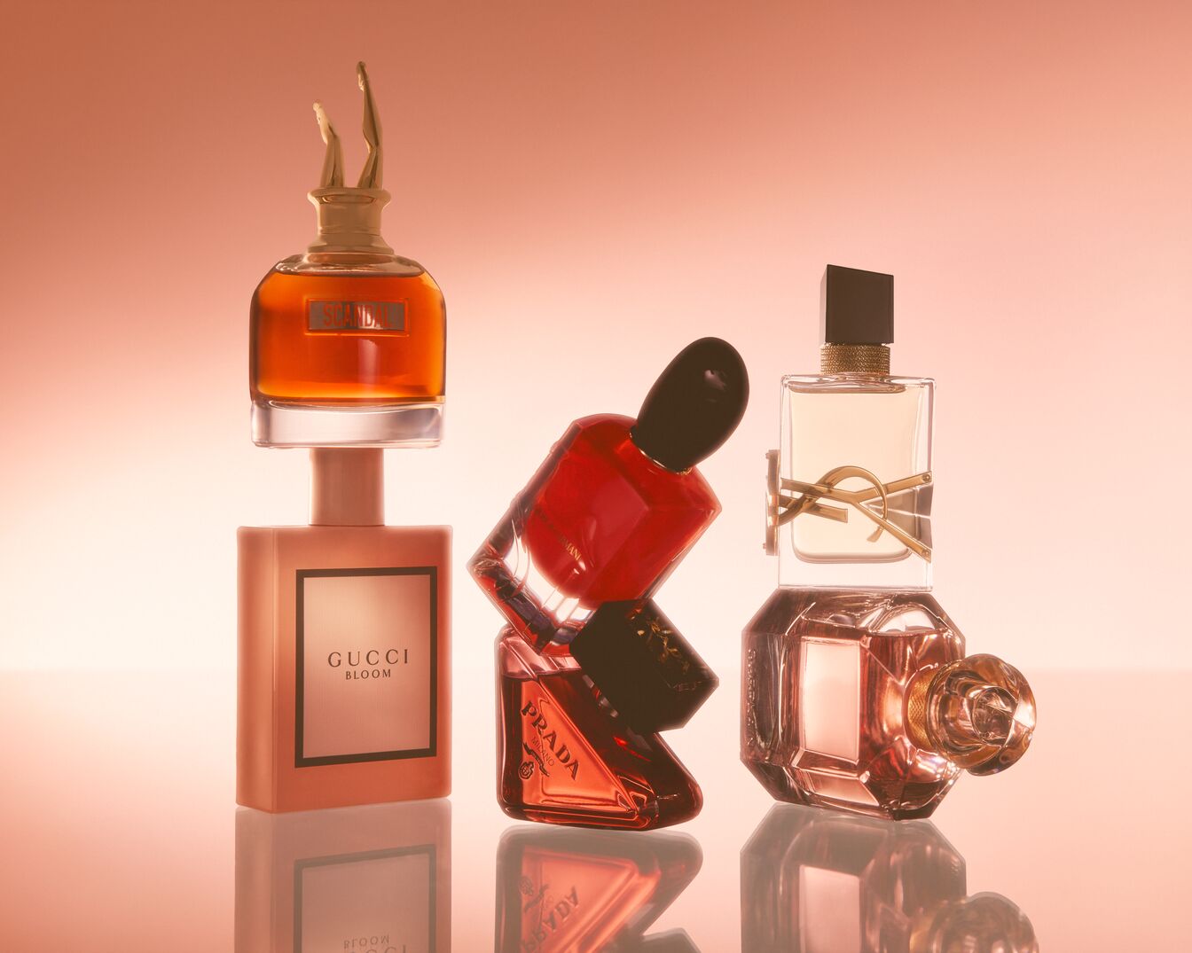 Šiame paveikslėlyje matyti kvepalų kolekcija iš tokių prekių ženklų kaip Gucci Bloom, Prada ir Yves Saint Laurent, išdėliota ant rausvo fono.