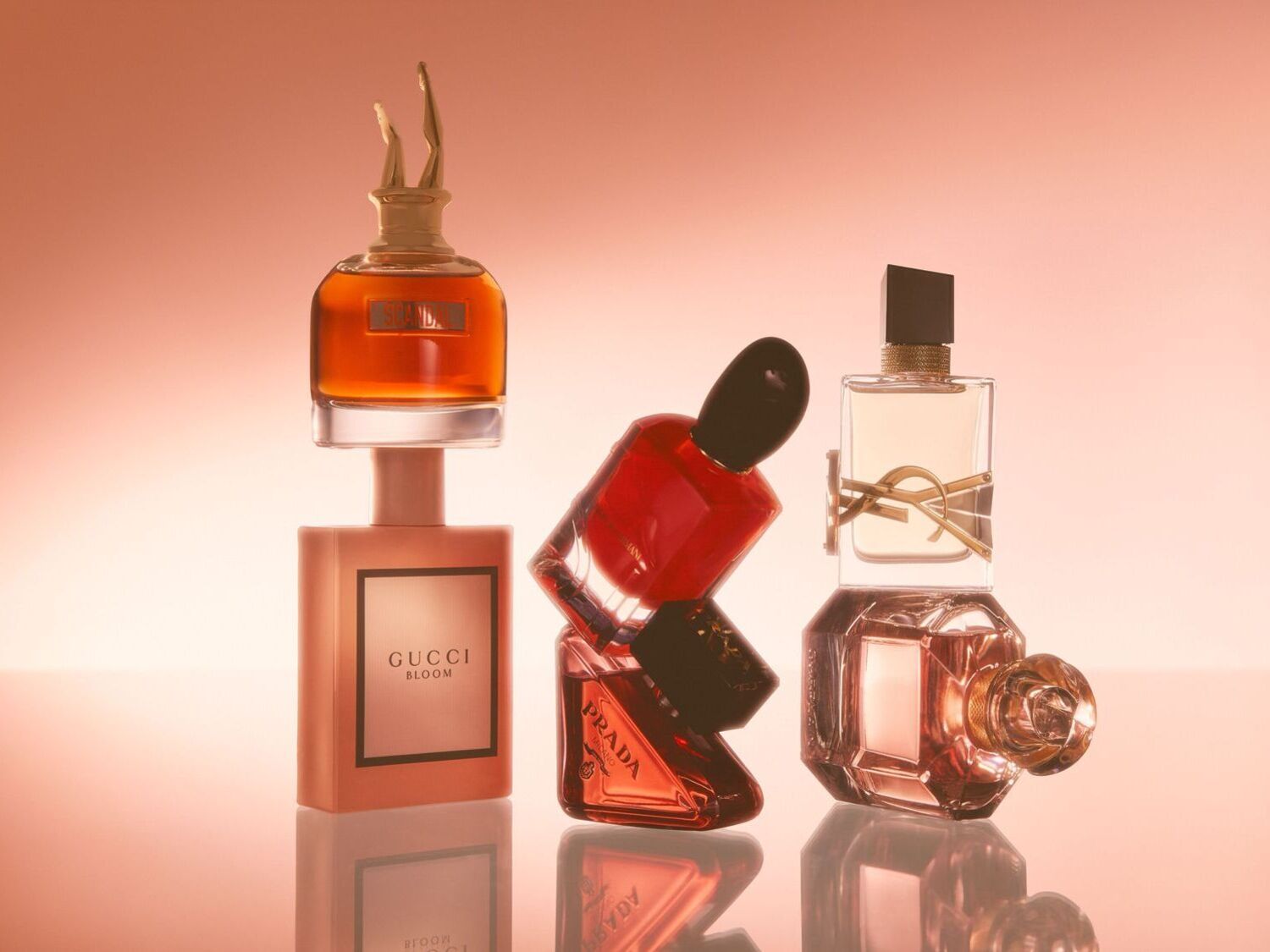 Įvairių kvepalų buteliukų kompozicija: Gucci Bloom švelniai rožinis, Prada raudonas, YSL Libre skaidrus su auksinėmis detalėmis ir Scandal rudas su auksinėmis kojomis.