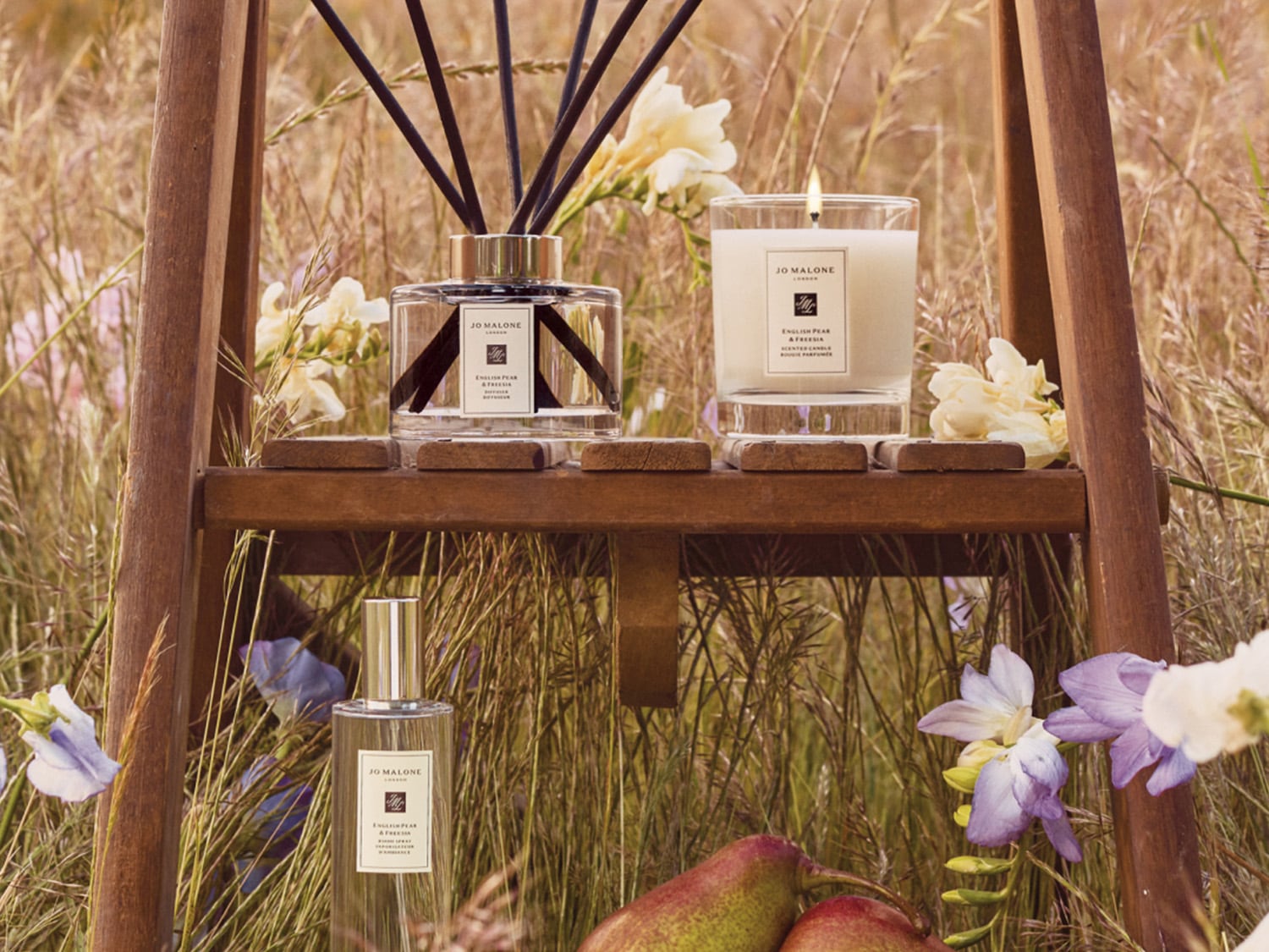 Auf einer Holzleiter in einer Blumenwiese sind Jo Malone Home-Produkte wie eine Duftkerze, ein Raumduft mit Duftstäbchen und ein Raumspray platziert.