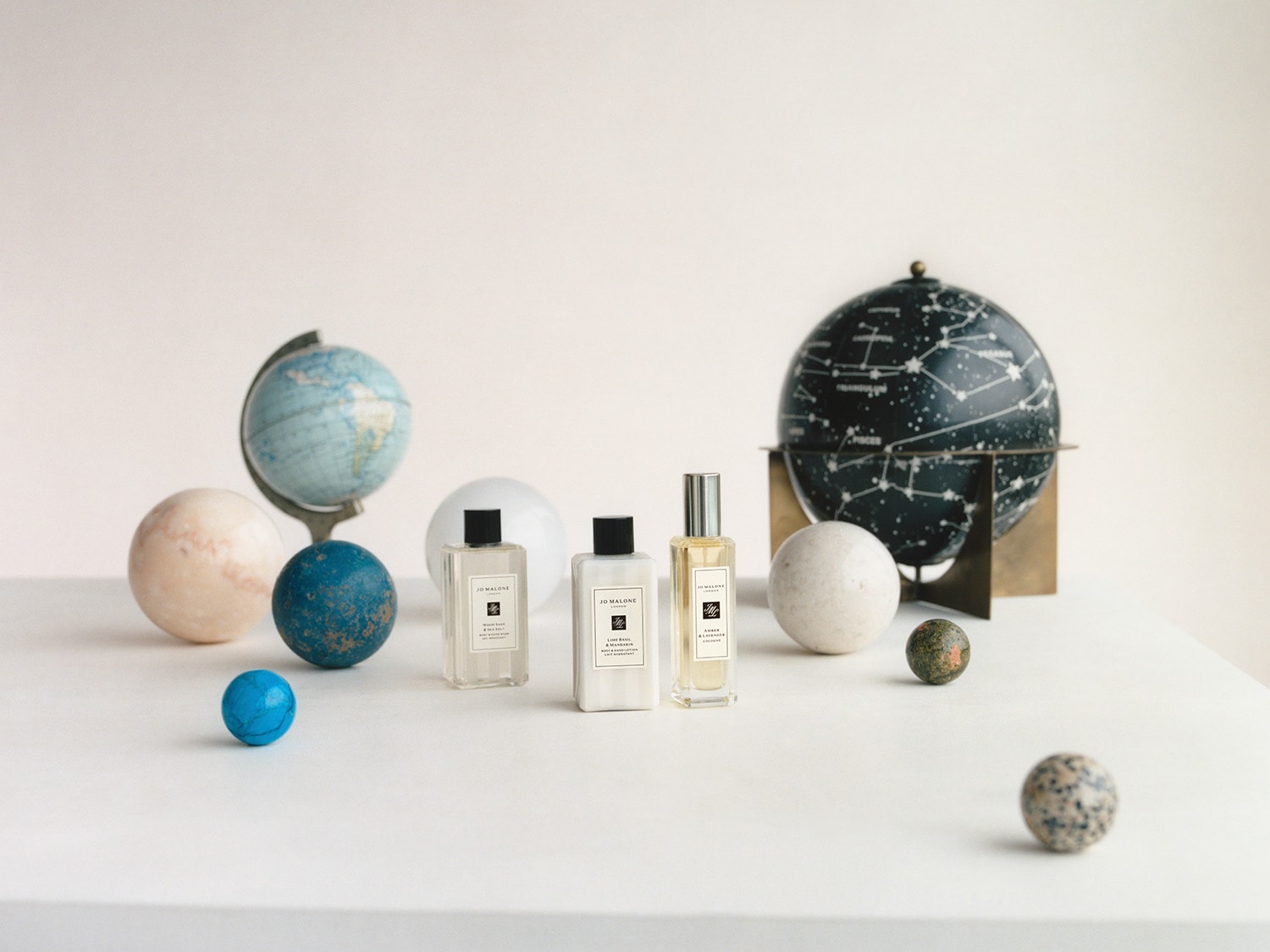 Auf einer weißen Oberfläche stehen Globusse und Kugeln in verschiedenen Größen neben drei Jo Malone Miniatur-Produkten.