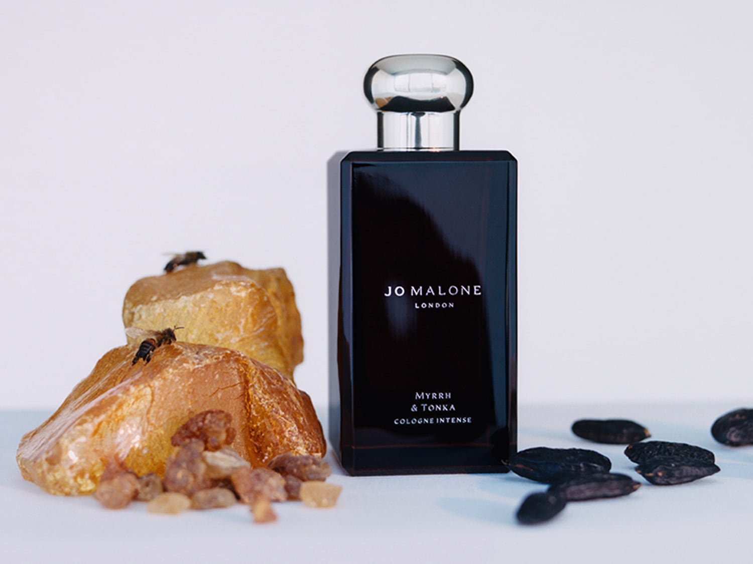 Jo Malone London Myrrh & Tonka Cologne Intense in einem dunklen, rechteckigen Flakon mit silbernem Verschluss, umgeben von goldenem Harz, Tonkabohnen und zwei Bienen.