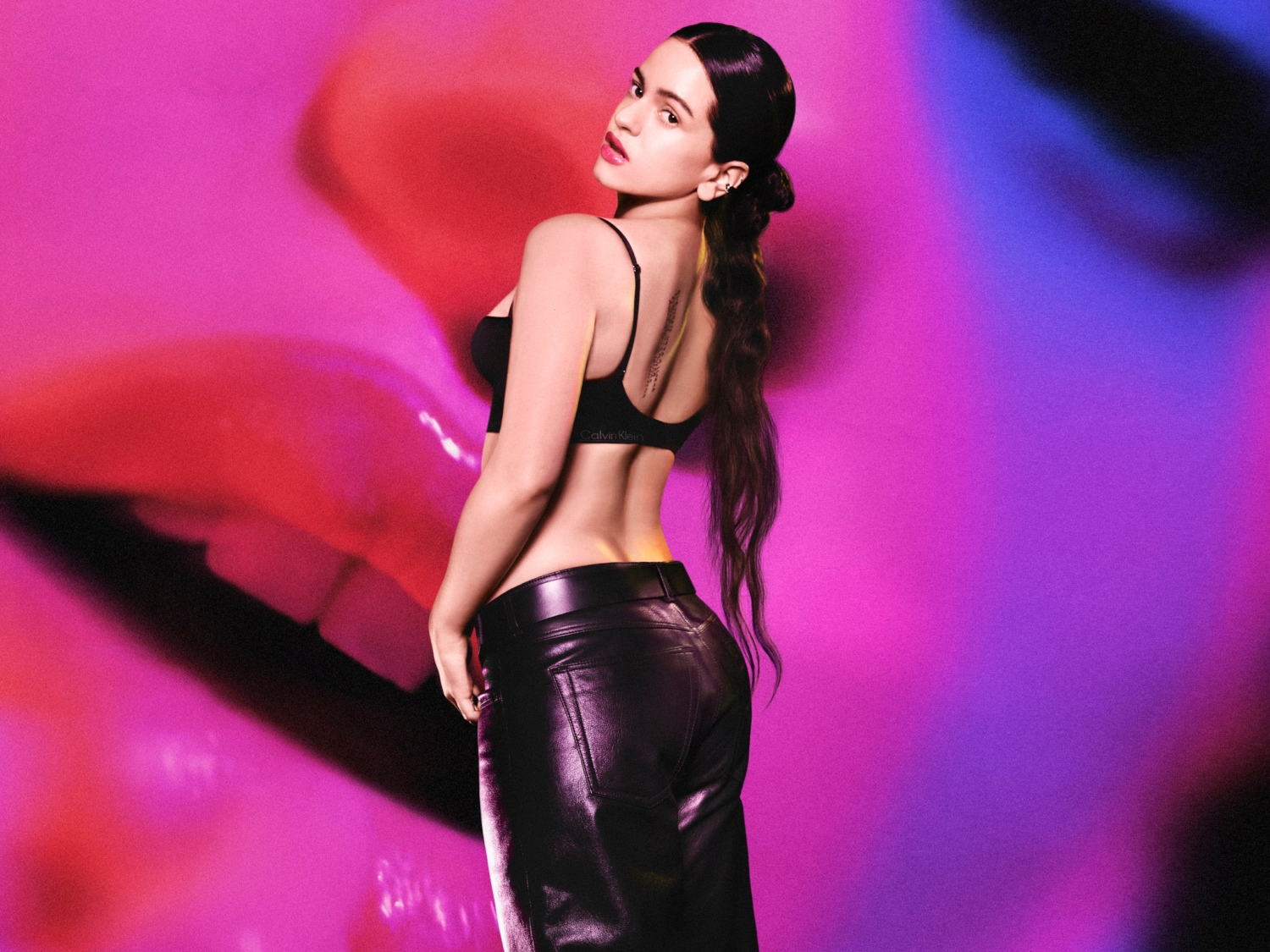 Kobieta w czarnym staniku Calvina Kleina i skórzanych spodniach pozuje na tle ust w odcieniach różu i fioletu.