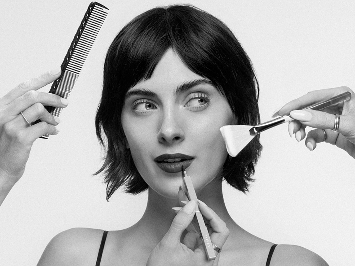 En la imagen en blanco y negro, una mujer de pelo corto se prepara con un peine, una brocha y un delineador de labios para un look impecable.