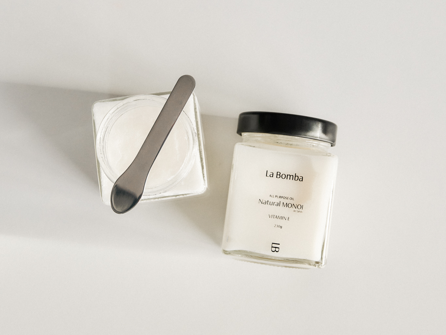 Na białym tle leżą dwa słoiki z olejem La Bomba Natural Monoi z witaminą E, jeden otwarty z metalową łyżką w środku, drugi zamknięty czarną zakrętką.