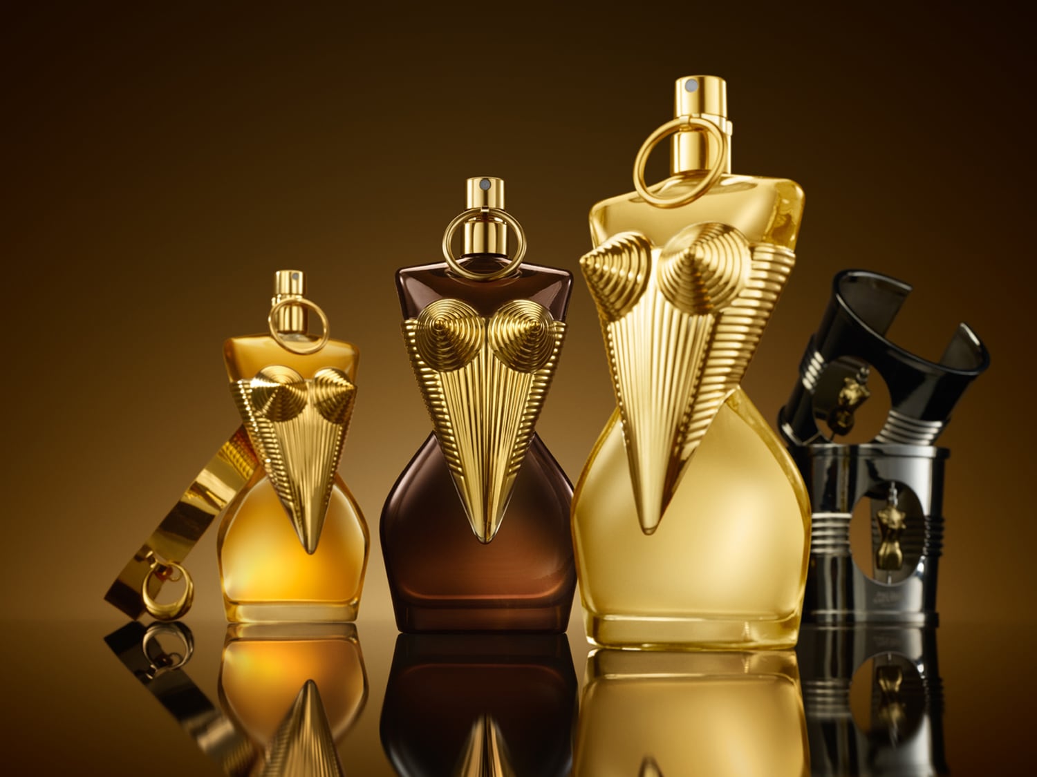 Trzy flakony perfum Jean Paul Gaultier La Belle w kształcie kobiecego torsu, w odcieniach złota i brązu, stojące na błyszczącej powierzchni.