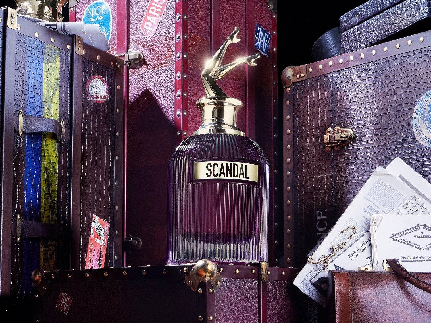 Fioletowy flakon perfum Scandal z korkiem w kształcie złotych nóg na tle skórzanych walizek z naklejkami z podróży, tworzący luksusową i tajemniczą kompozycję.