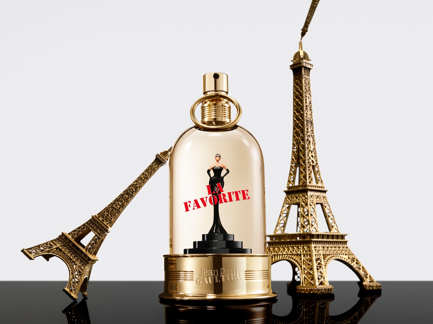 Złoty flakon perfum Jean Paul Gaultier z figurką kobiety w czarnej sukience i napisem "I FAVORITE" w otoczeniu dwóch złotych wież Eiffla, stojący na czarnej, błyszczącej powierzchni.