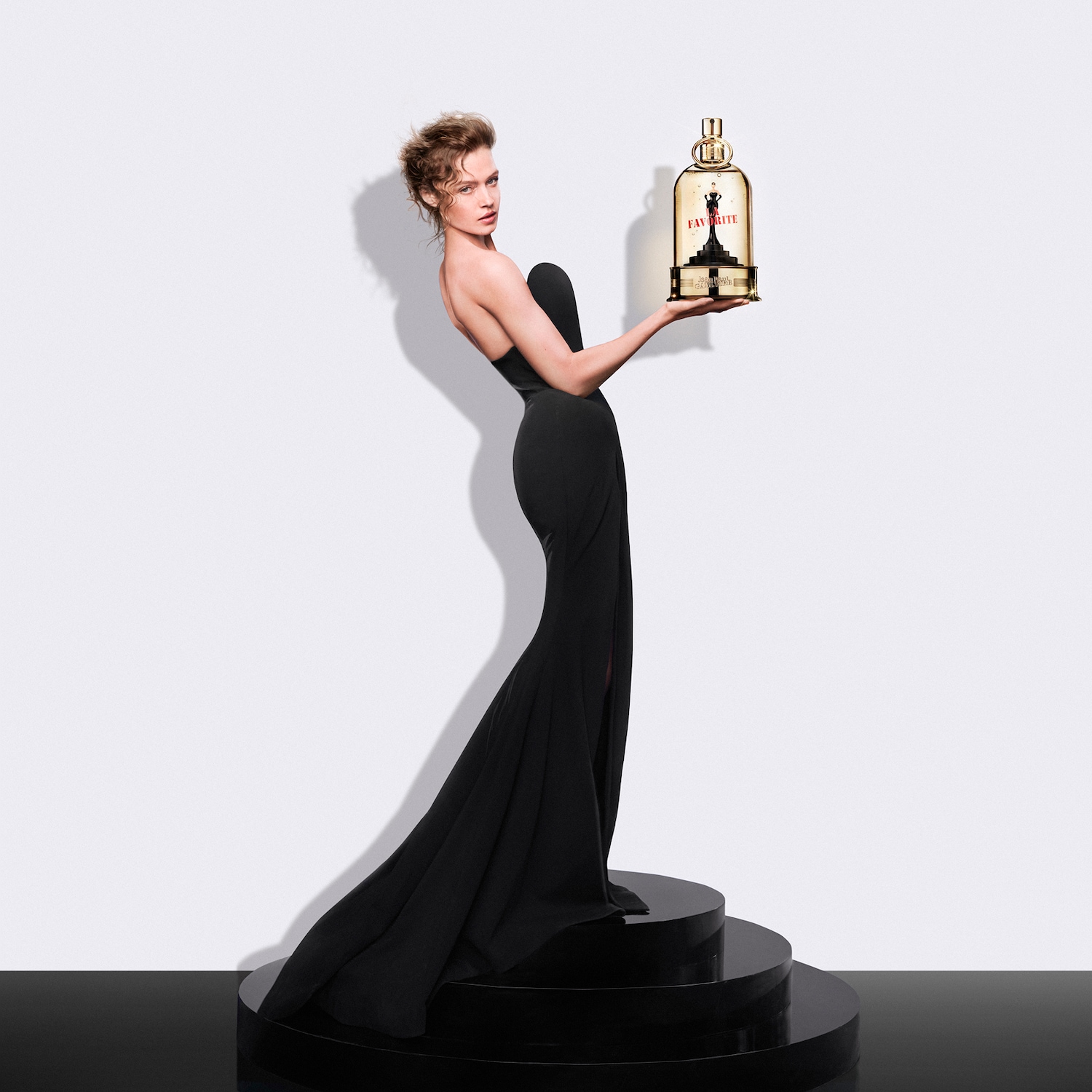Modelka w czarnej sukni bez ramiączek prezentuje flakon perfum "Favorite" w kształcie złotej klatki z figurką w środku, stojąc na czarnym, okrągłym cokole.