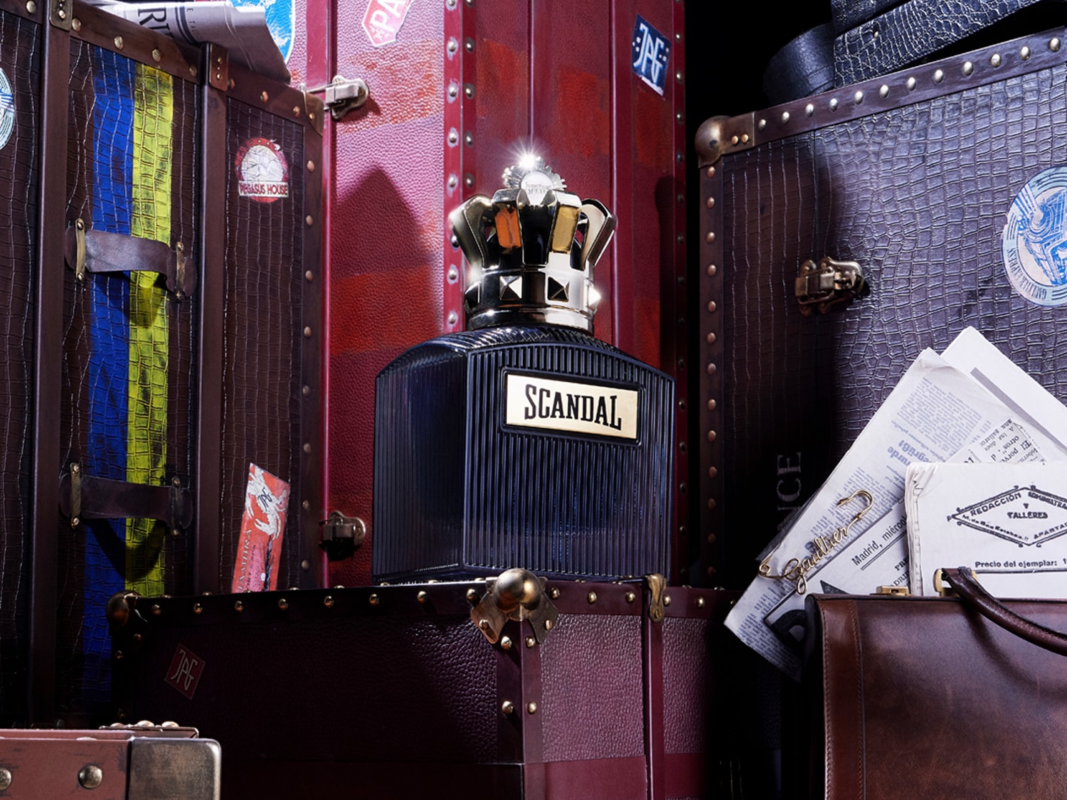 Flakon perfum Scandal z czarnymi pionowymi paskami i złotą koroną na tle skórzanych walizek w stylu vintage.