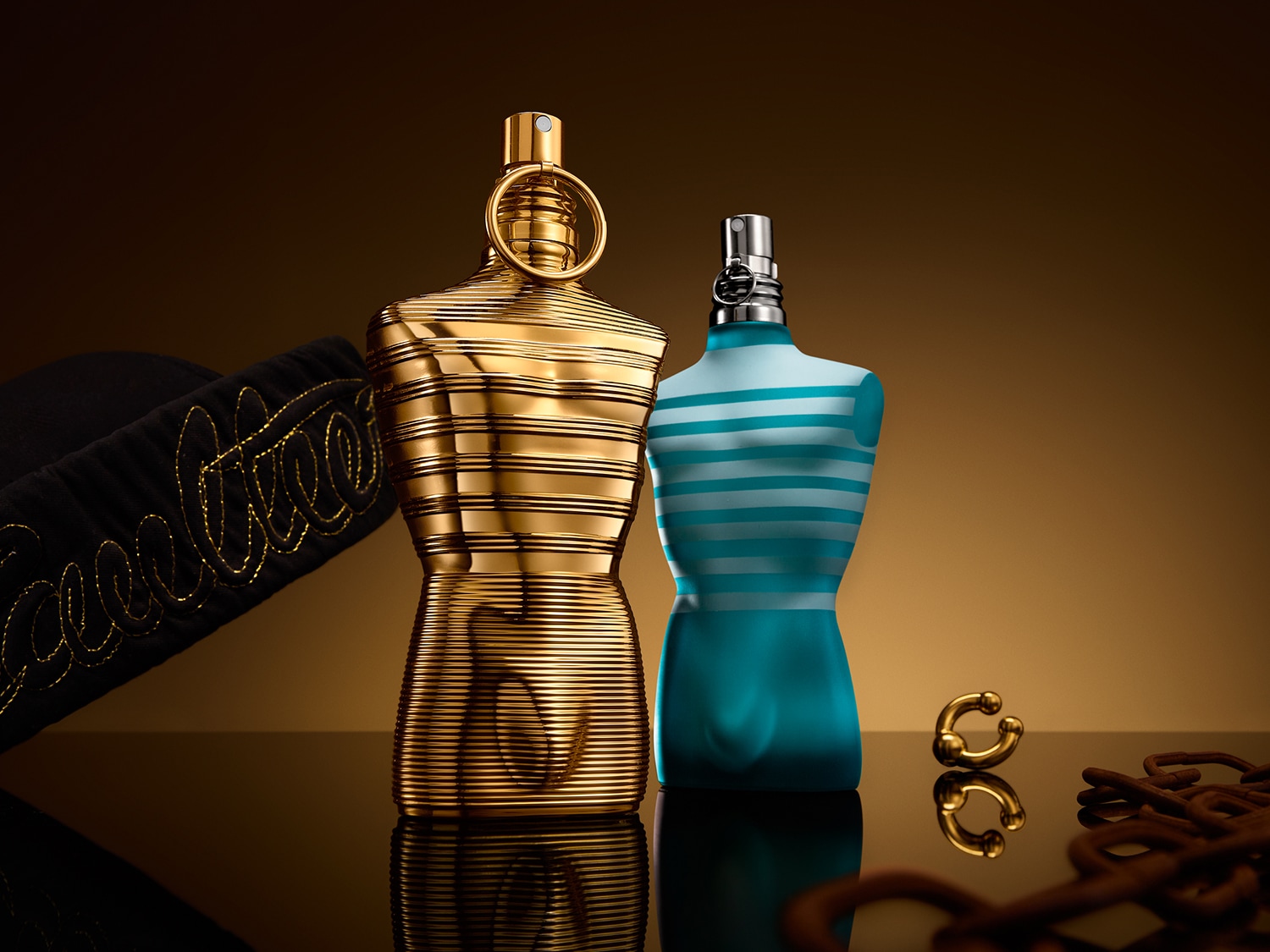 Dwa flakony perfum w kształcie męskiego torsu: jeden złoty, prążkowany, drugi w błękitne paski, stoją obok czapki i złotego kolczyka na tle bursztynowego światła.