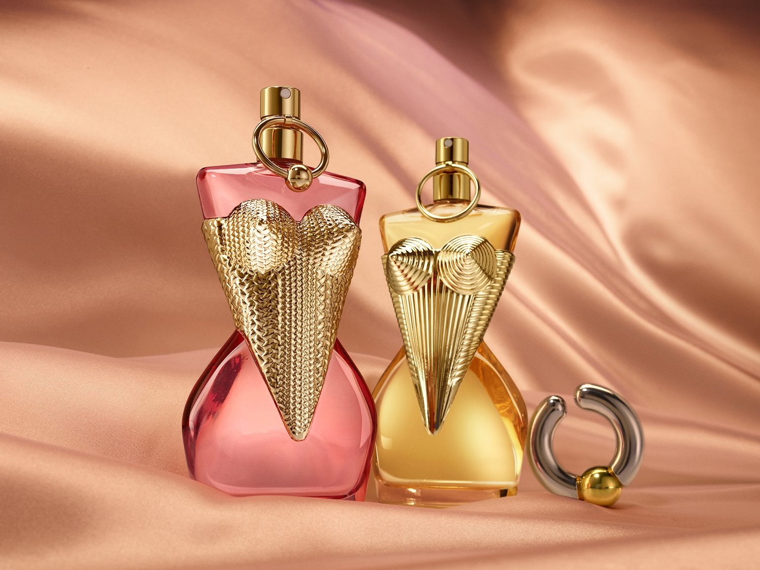Dwa flakony perfum w kształcie kobiecej sylwetki, jeden w kolorze różowym, a drugi w kolorze złotym, stoją na jedwabistym, brzoskwiniowym tle, emanując luksusem i wyrafinowaniem.
